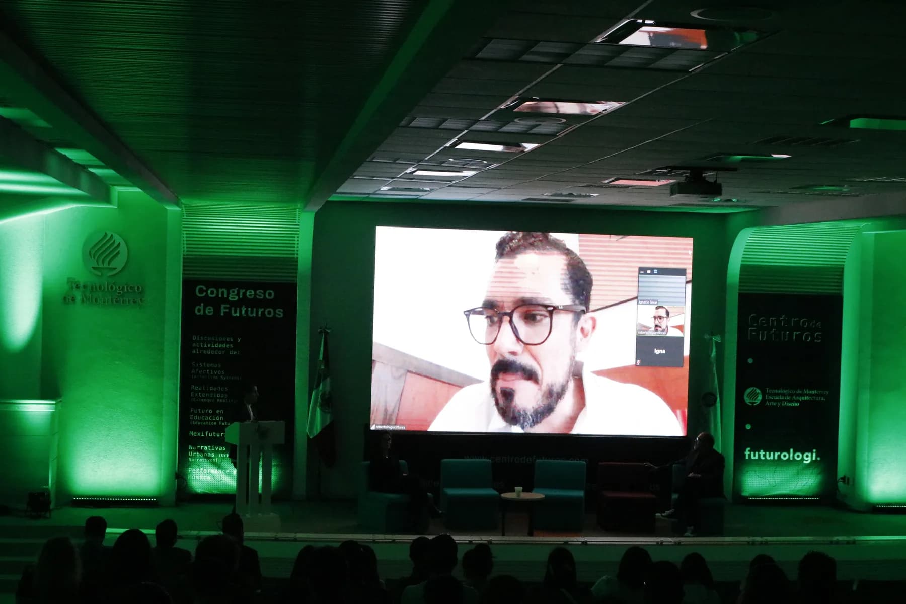 Escenario iluminado en verde con panelistas y una presentación remota ante el público